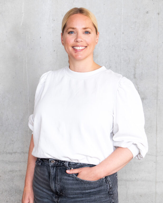 En person med blondt hår sat op i en lav knold står og smiler, iført en hvid langærmet skjorte og mørke jeans, med ryggen mod en lysegrå betonvæg og den ene hånd i lommen.