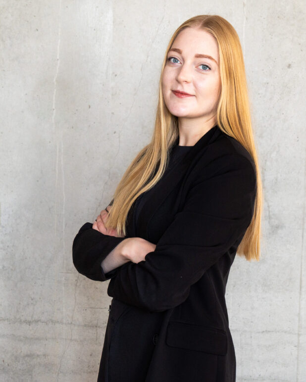 En kvinde med langt blondt hår, iført en sort blazer, står med armene over kors og smiler let foran en enkel, lys betonvæg.